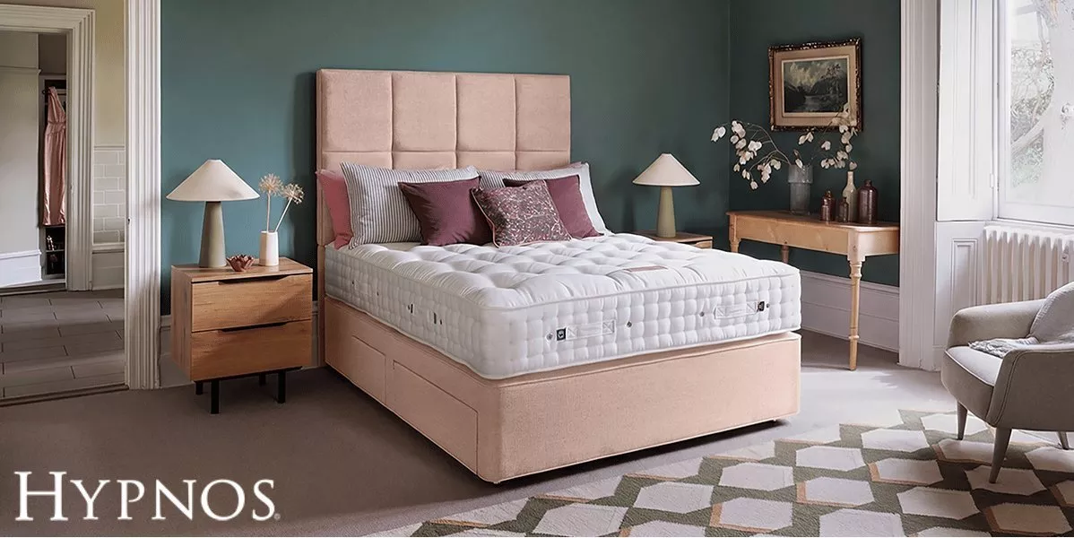 A plush tufted mattress on a light pink upholstered divan bed, styled with plum pillows in a bedroom with teal walls, wooden nightstands, matching table lamps, and a console table.